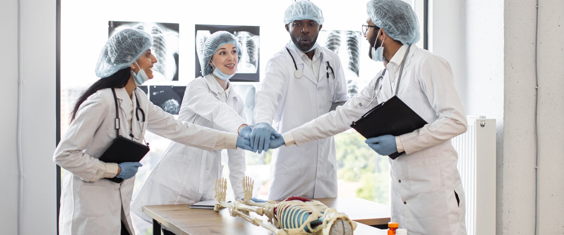 Multinational conference of doctors in a modern clinic. Group of successful medics celebrating success by touching hands as team sign together at table with human skeleton model.